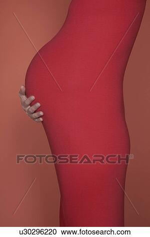 Stock Image - Pregnant woman in body suit holding abdomen, mid section, side view, studio shot. Fotosearch