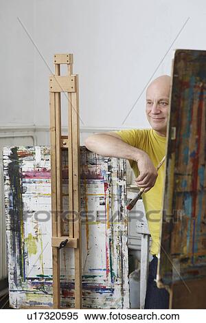 Stock Photography - Relaxed male artist with paint brush leaning against easel. Fotosearch