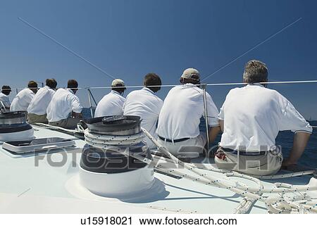 Stock Image - Sailing crew sitting on side of yacht on ocean, back view. Fotosearch