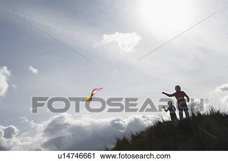 Stock Image - Silhouette of girl and father flying kite on dunes. Fotosearch