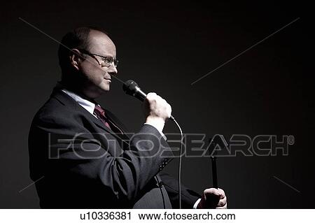 Stock Image - Smiling man in full suit holding microphone, side view. Fotosearch
