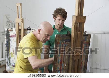 Stock Image - Student looking on as artist paints at easel in art studio. Fotosearch