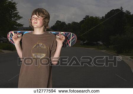 Teenage boy (13-15) holding skateboard standing on street View Large Photo Image Picture - Teenage boy (13-15) holding skateboard standing on street. Fotosearch