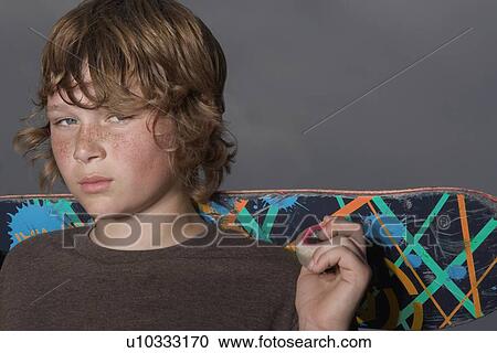 Stock Image - Teenage boy (13-15) holding skateboard, outdoors, portrait, close up. Fotosearch
