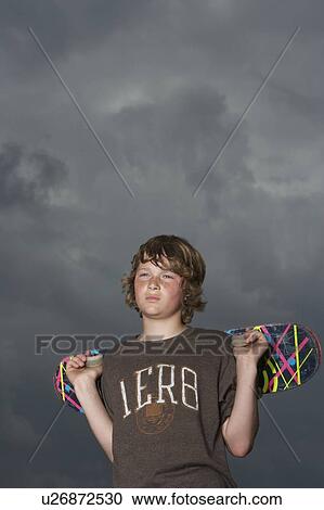 Stock Image - Teenage boy (13-15) holding skateboard, standing against sky, low angle view. Fotosearch