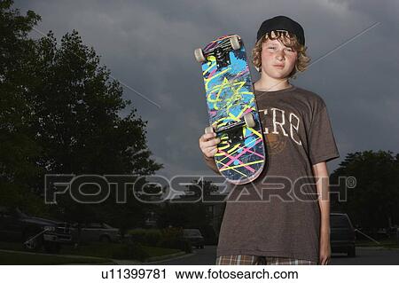 Stock Image - Teenage boy holding skateboard, standing on street, low angle view, portrait. Fotosearch