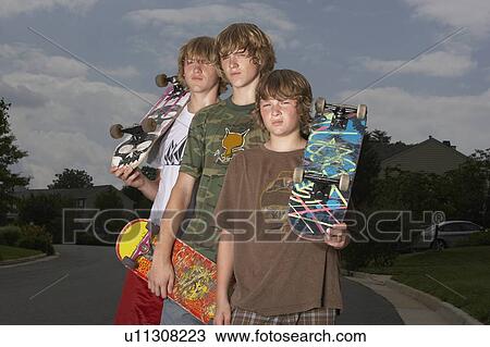 Stock Image - Three teenage brothers (13-17) standing on street holding skateboards, portrait. Fotosearch