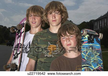 Stock Image - Three teenage brothers (13-17) standing on street holding skateboards, portrait. Fotosearch