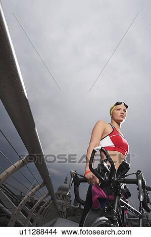 Picture - Triathlon athlete holding bike standing on footbridge, low angle view, Millennium Bridge, London, England. Fotosearch