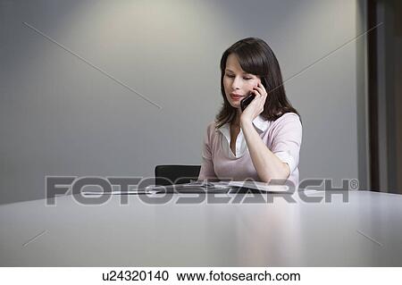 Stock Image - Woman sitting at desk, talking on mobile phone. Fotosearch