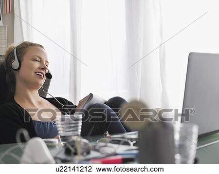 Stock Image - Woman using laptop wearing headset sitting at desk. Fotosearch