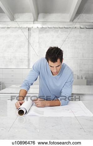 Young man leaning on table, looking at blueprint View Large Photo Image Stock Photograph - Young man leaning on table, looking at blueprint. Fotosearch