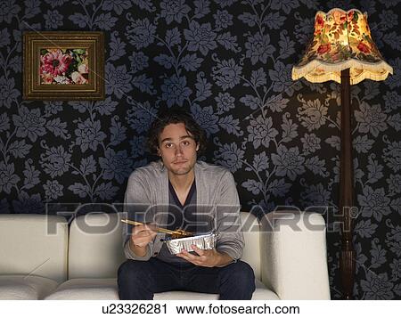 Young man sitting on sofa holding take out food View Large Photo Image Stock Image - Young man sitting on sofa holding take out food. Fotosearch
