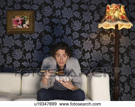 Young man sitting on sofa holding take out food View Large Photo Image Stock Photo - Young man sitting on sofa holding take out food. Fotosearch