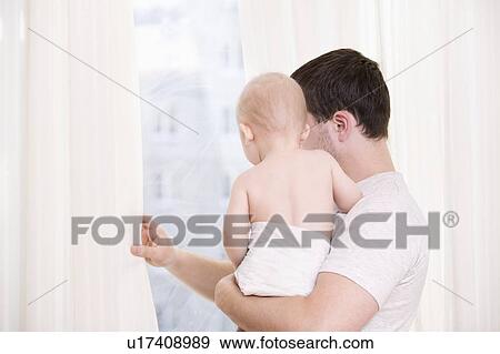 Stock Photo - Young man stands looking through curtains with toddler son. Fotosearch