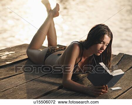 Stock Image - Young woman in bikini lying on jetty reading book. Fotosearch