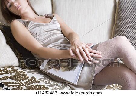 Stock Image - Young woman in silver dress sits on sofa holding magazine. Fotosearch