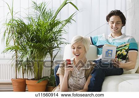 Picture - Young woman listening mp3 player next to man reading book on sofa. Fotosearch