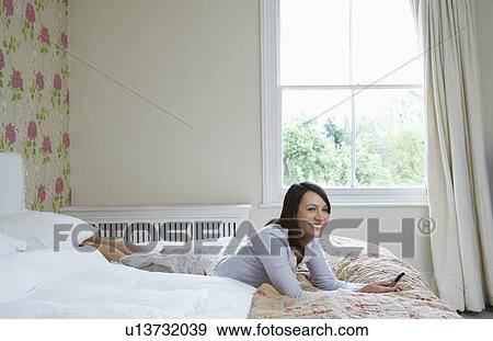 Young woman lying on bed holding mobile phone, smiling View Large Photo Image Stock Photo - Young woman lying on bed holding mobile phone, smiling. Fotosearch