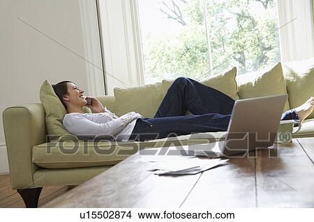 Picture - Young woman lying on sofa while talking on mobile. Fotosearch