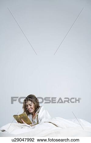 Stock Image - Young woman reading book in bed, smiling. Fotosearch