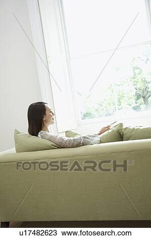 Stock Image - Young woman relaxing on sofa by window. Fotosearch