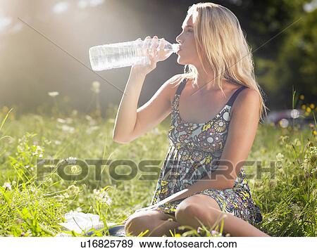 Stock Photo - Young woman sitting in meadow drinking water from bottle. Fotosearch