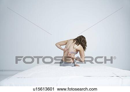 Stock Image - Young woman sitting on bed, fastening bra, back view. Fotosearch