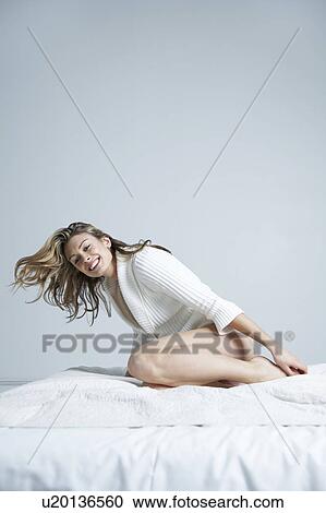Stock Image - Young woman sitting on bed, smiling, portrait. Fotosearch