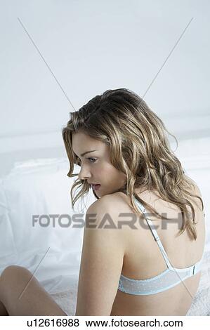 Stock Photo - Young woman wearing bra sitting on bed, elevated view. Fotosearch