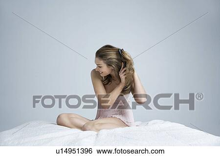 Stock Photograph - Young woman wearing camisole, brushing hair on bed. Fotosearch