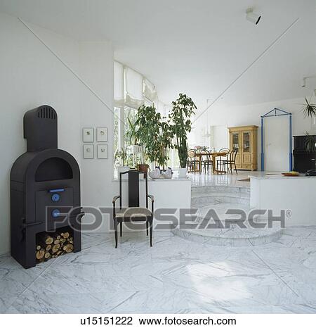 Stock Image - Antique chair beside black cast iron stove in openplan split-level hall dining room with marble floor tiles. Fotosearch