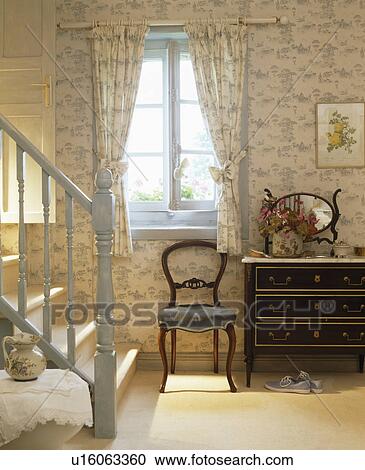 Stock Image - Antique chair in front of window with blue+cream curtains in country bedroom with pale blue painted stairs. Fotosearch