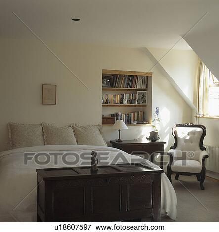 Antique chest below bed with cream bedlinen in attic bedroom with alcove shelving and white armchair View Large Photo Image Stock Photo - Antique chest below bed with cream bedlinen in attic bedroom with alcove shelving and white armchair. Fotosearch