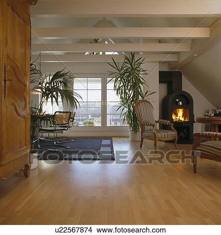 Picture - Black cast-iron stove with lighted fire in modern openplan living room with tall houseplants and wooden flooring. Fotosearch