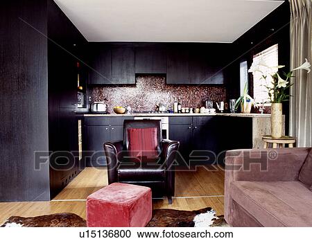 Stock Image - Black leather armchair and pale brown suede sofa in open-plan kitchen and living room in modern apartment. Fotosearch