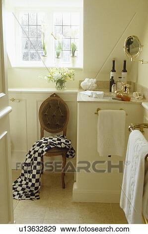 Stock Photo - Black+white chequerboard towel on antique chair beside basin in vanity unit in cream bathroom. Fotosearch