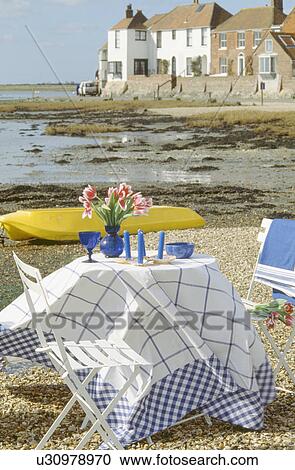 Stock Image - Blue glasses and blue+white checked cloth on table on beach in small harbour. Fotosearch