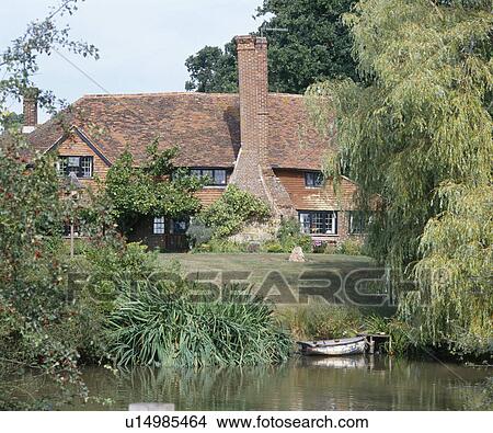 Picture - Boat moored on river bank with large willow trees in front of lawn and old red brick house with tall chimney. Fotosearch
