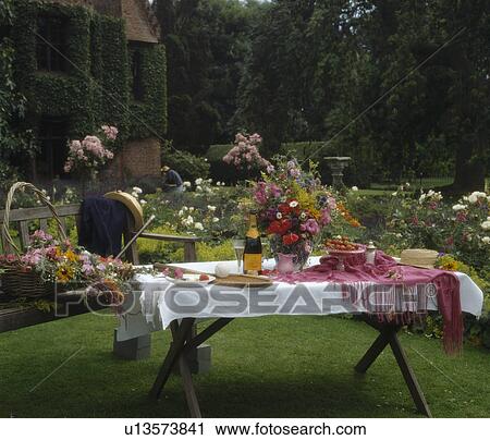 Stock Image - Bottle of champagne and white linen cloth on table in country garden in summer. Fotosearch