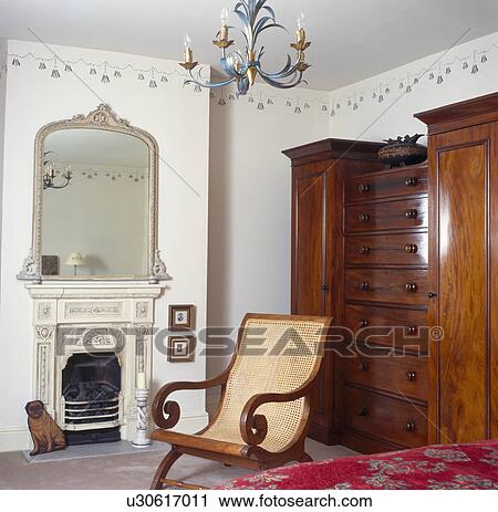 Cane-backed planter's chair and large mahogany wardrobe in bedroom with painted mirror above ornate white fireplace View Large Photo Image Stock Image - Cane-backed planter's chair and large mahogany wardrobe in bedroom with painted mirror above ornate white fireplace. Fotosearch