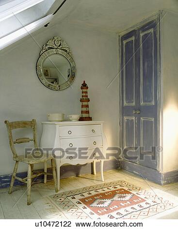 Stock Image - Chair beside white chest-of-drawers in attic bedroom with blue door. Fotosearch