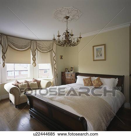 Chandelier above lit bateau bed with cream bedlinen in townhouse bedroom with cream blind on bay window View Large Photo Image Stock Photography - Chandelier above lit bateau bed with cream bedlinen in townhouse bedroom with cream blind on bay window. Fotosearch