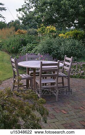 Stock Image - Circular wooden table and chairs on herringbone brick paved patio in summer country garden. Fotosearch
