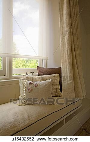 Stock Image - Close-up of cream cushions on windowseat below white blind on window. Fotosearch