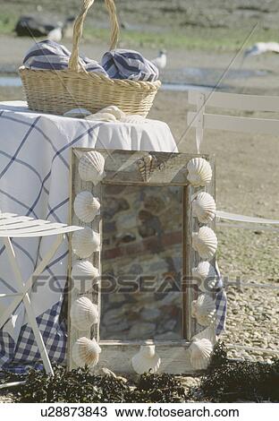 Stock Image - Close-up of mirror with seashell-decorated frame beside table with checked cloth. Fotosearch