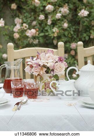 Stock Photograph - Close-up of pale pink roses in jug on table set for tea with white cloth and teaset and glasses of fruit juice. Fotosearch