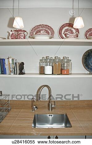 Stock Photography - Close-up of shelves above chrome mixer tap and small underset stainless-steel sink in apartment kitchen. Fotosearch