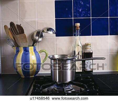 Stock Photograph - Close-up of utensils in blue striped jug beside steel pan on gas hob. Fotosearch