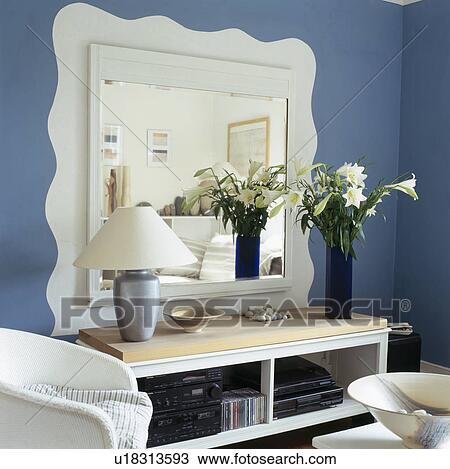 Stock Image - Close-up of white mirror above console table with lamp and flower arrangement. Fotosearch